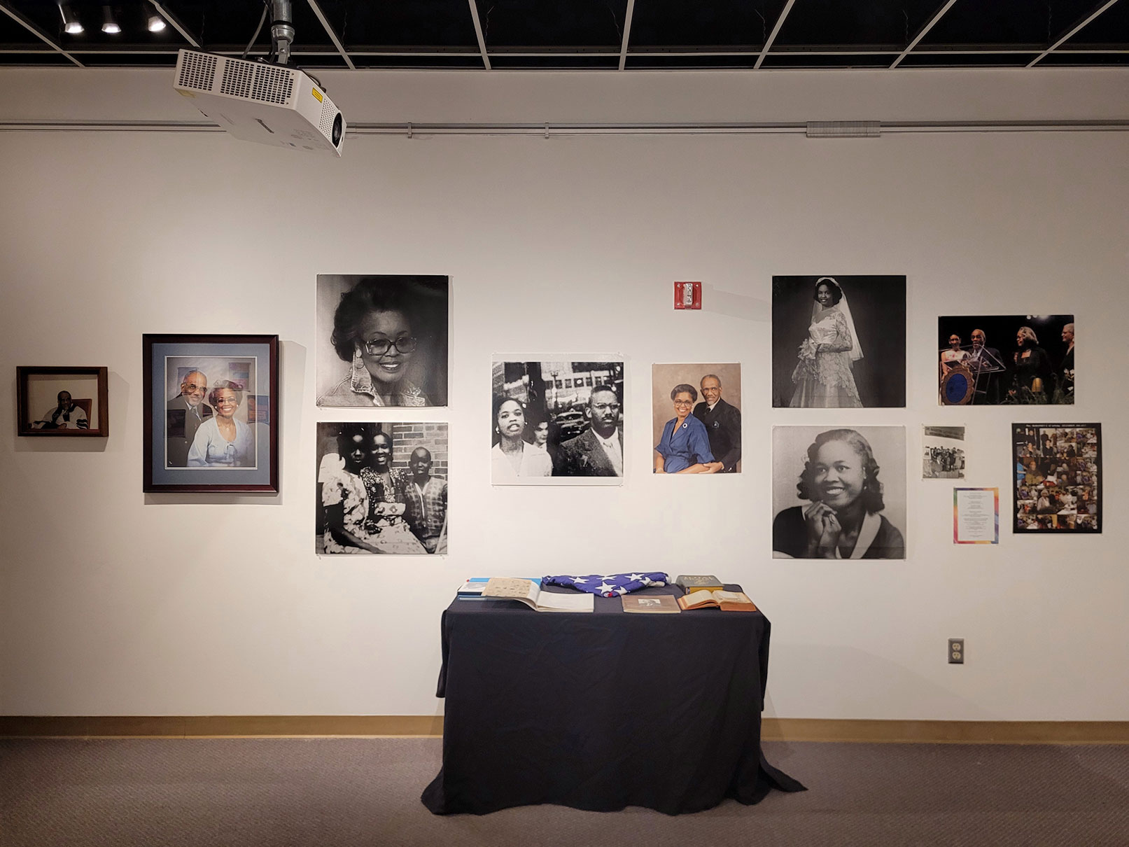 A photograph of an art installation with photos of Dr. Samuel B. McKinney’s wife and family on the wall behind a table draped in a black tablecloth with various books, an open bible with ephemera in the pages, and a ceremonial coffin flag.